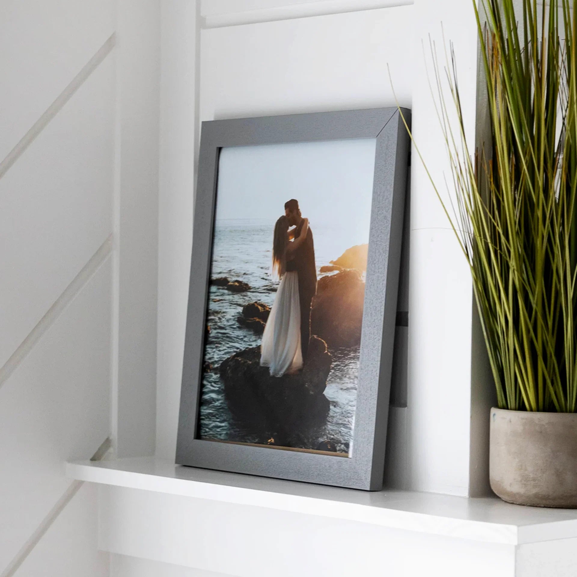 Gray framed vertical wedding photo of a couple kissing on ocean rocks at sunset, displayed on a white shelf beside a tall plant.