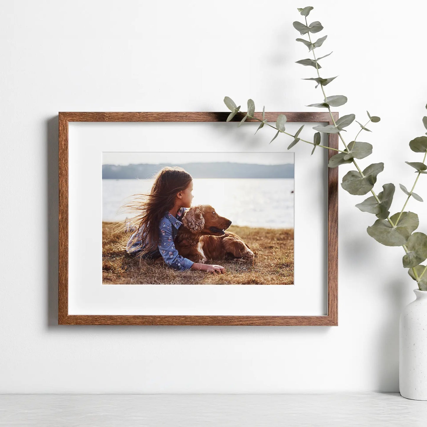 Framed photo print showing a child lying beside a dog near a lake, displayed on a wall with greenery.