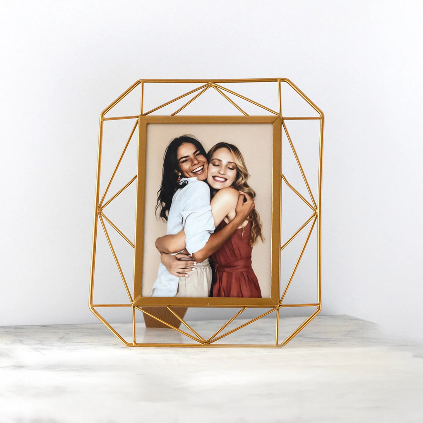 Tabletop photo print in a gold geometric frame showing two people hugging and smiling, displayed on a light surface
