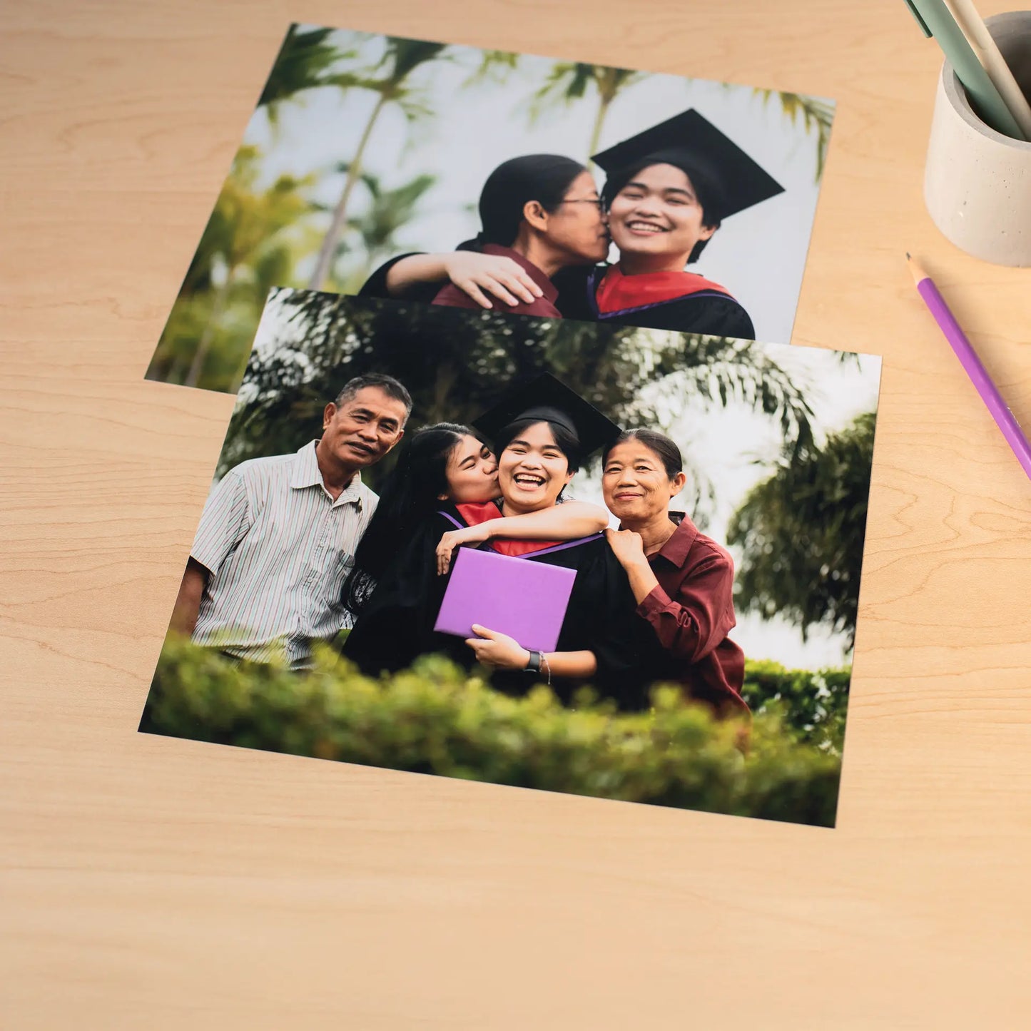 Two standard photo prints showing a graduate with family members outdoors, placed on a wooden desk with a pencil nearby.