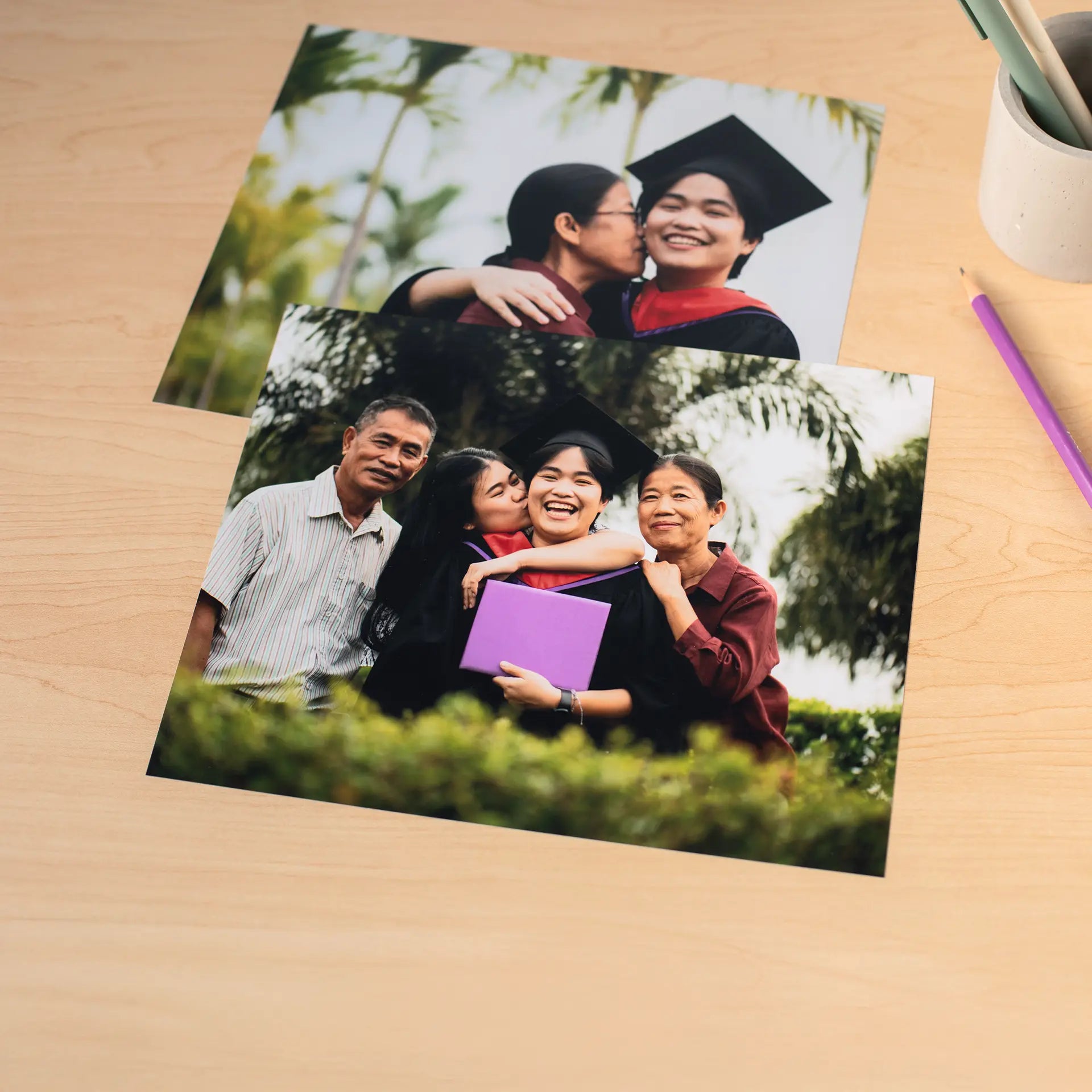 Two standard photo prints showing a graduate with family members outdoors, placed on a wooden desk with a pencil nearby.