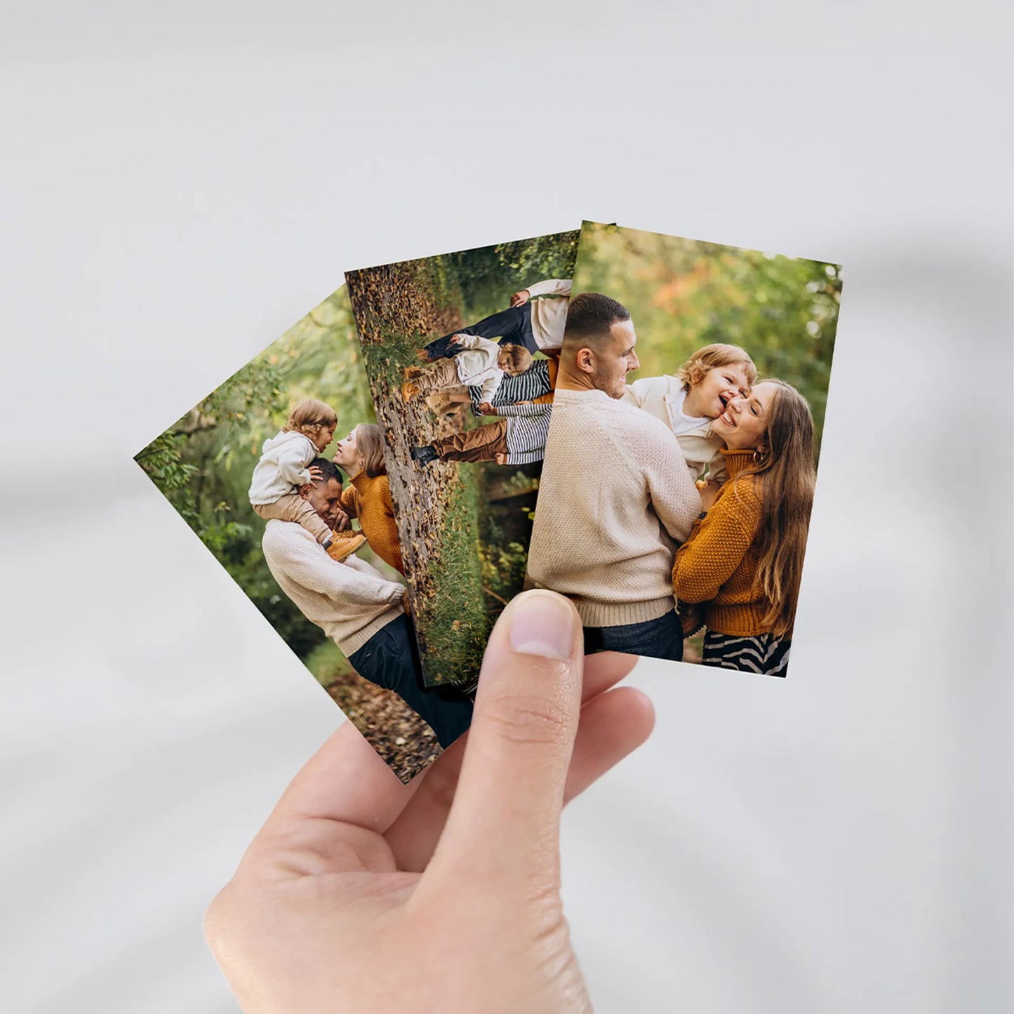 Hand holding two wallet sized photo prints featuring a family with a child outdoors in a leafy setting.