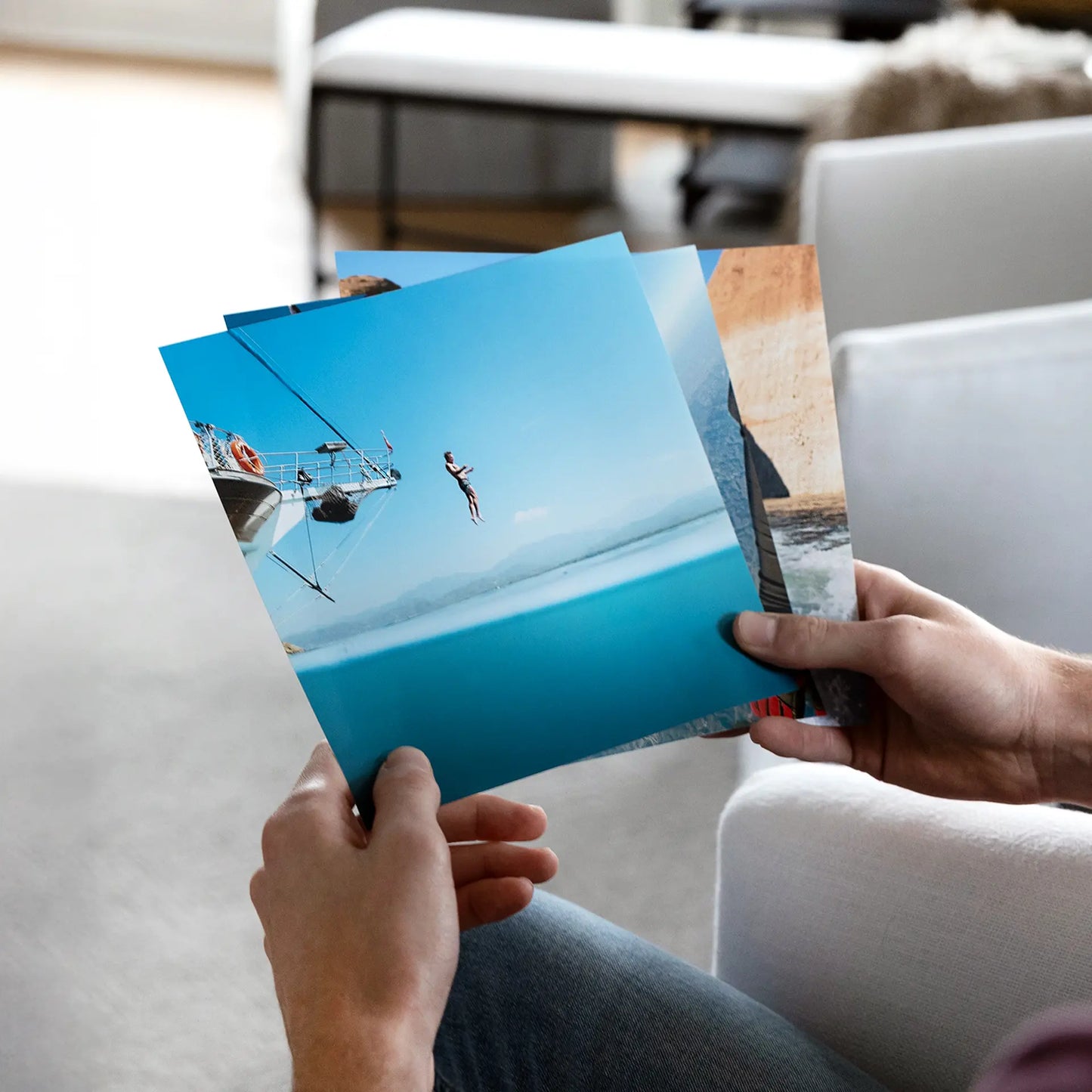 Hands holding multiple glossy photo prints featuring a person jumping into blue water, viewed in a casual indoor setting.