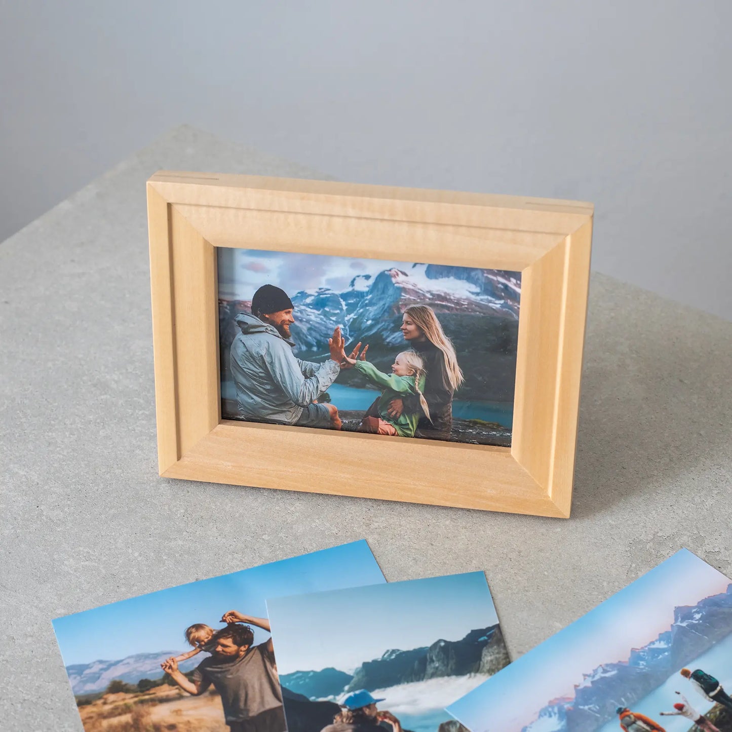 Small horizontal photo print in a light wood frame showing a family with a child outdoors in a mountain setting, placed on a tabletop.