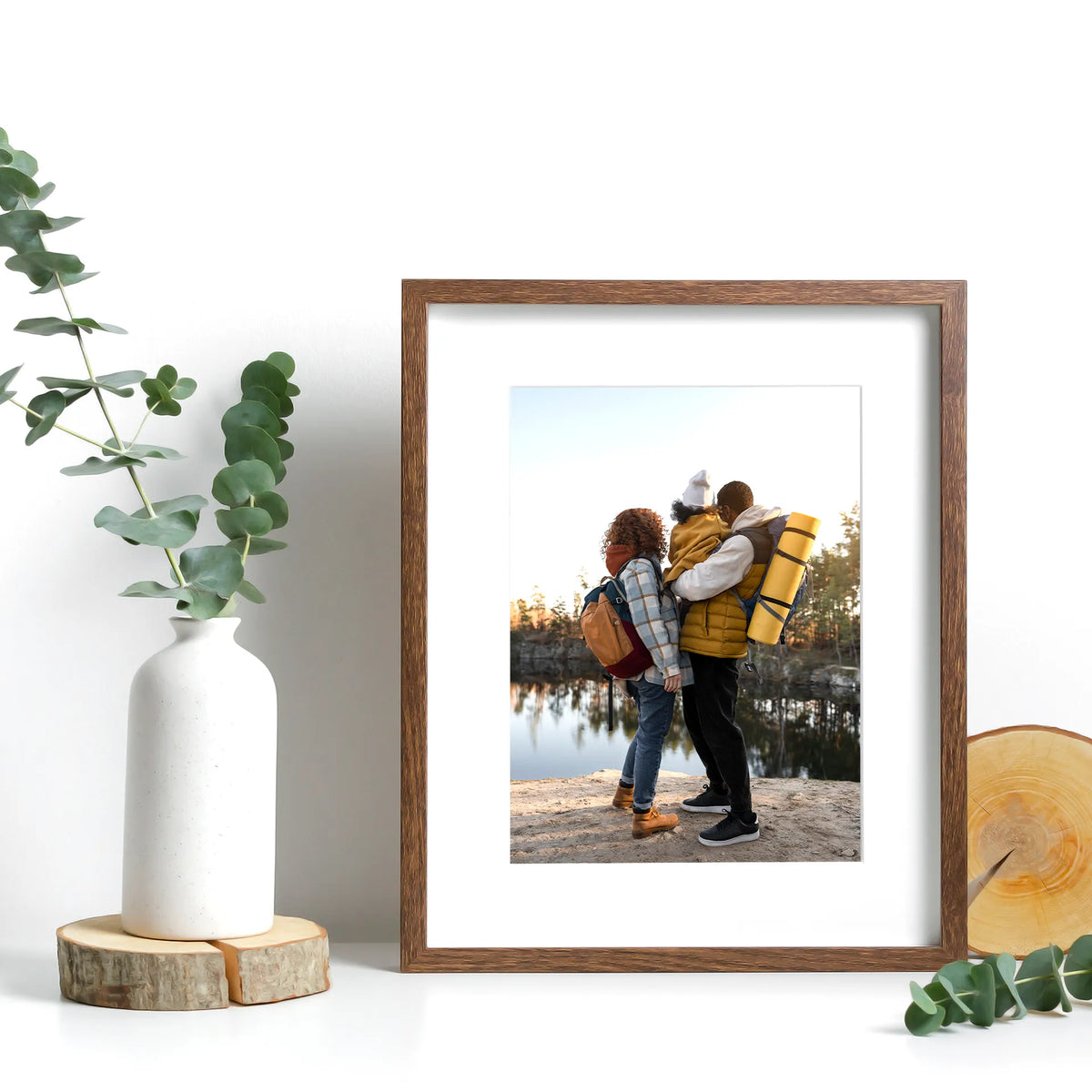 Standard print in a wood frame with white matting, showing a family with backpacks looking out over a lake.