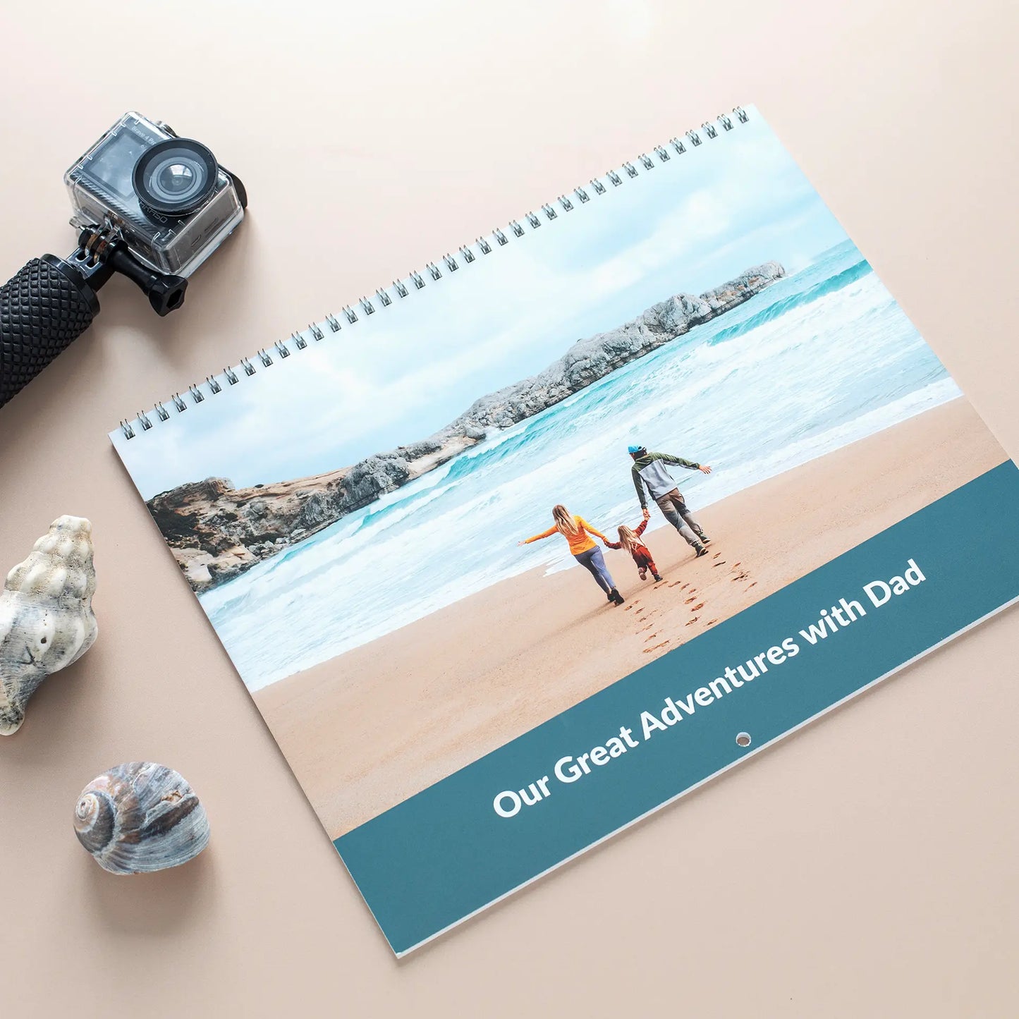 Wall calendar cover with beach scene of family walking along shore, text reads “Our Great Adventures with Dad,” placed beside camera and shells.