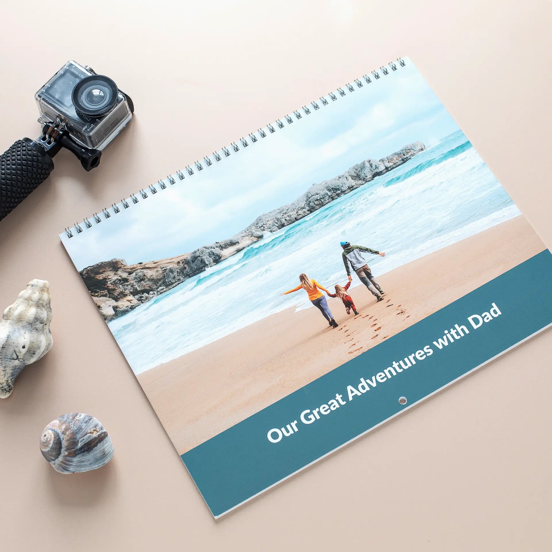 Wall calendar cover with beach scene of family walking along shore, text reads “Our Great Adventures with Dad,” placed beside camera and shells.