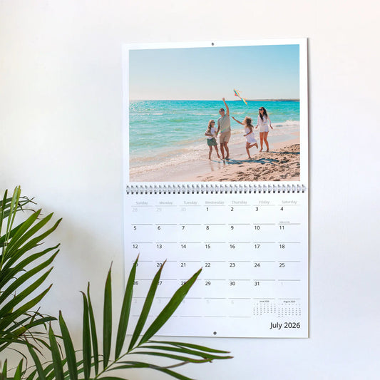 A wall calendar displayed on a white wall featuring a bright summer photo of a family of four laughing and flying a kite on a sunny beach, showing the month of July 2026.