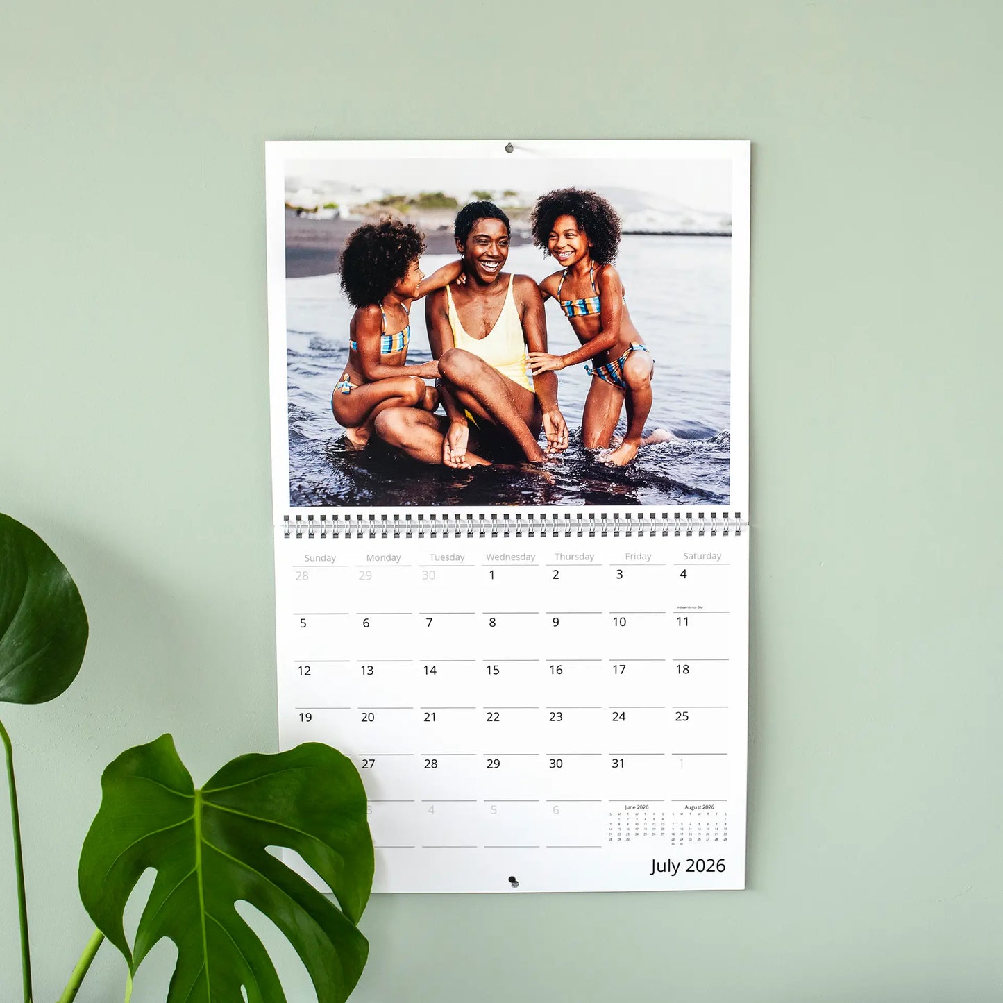 Wall calendar open to July 2026 featuring adult and two children sitting in shallow water, mounted on green wall with plant leaf.