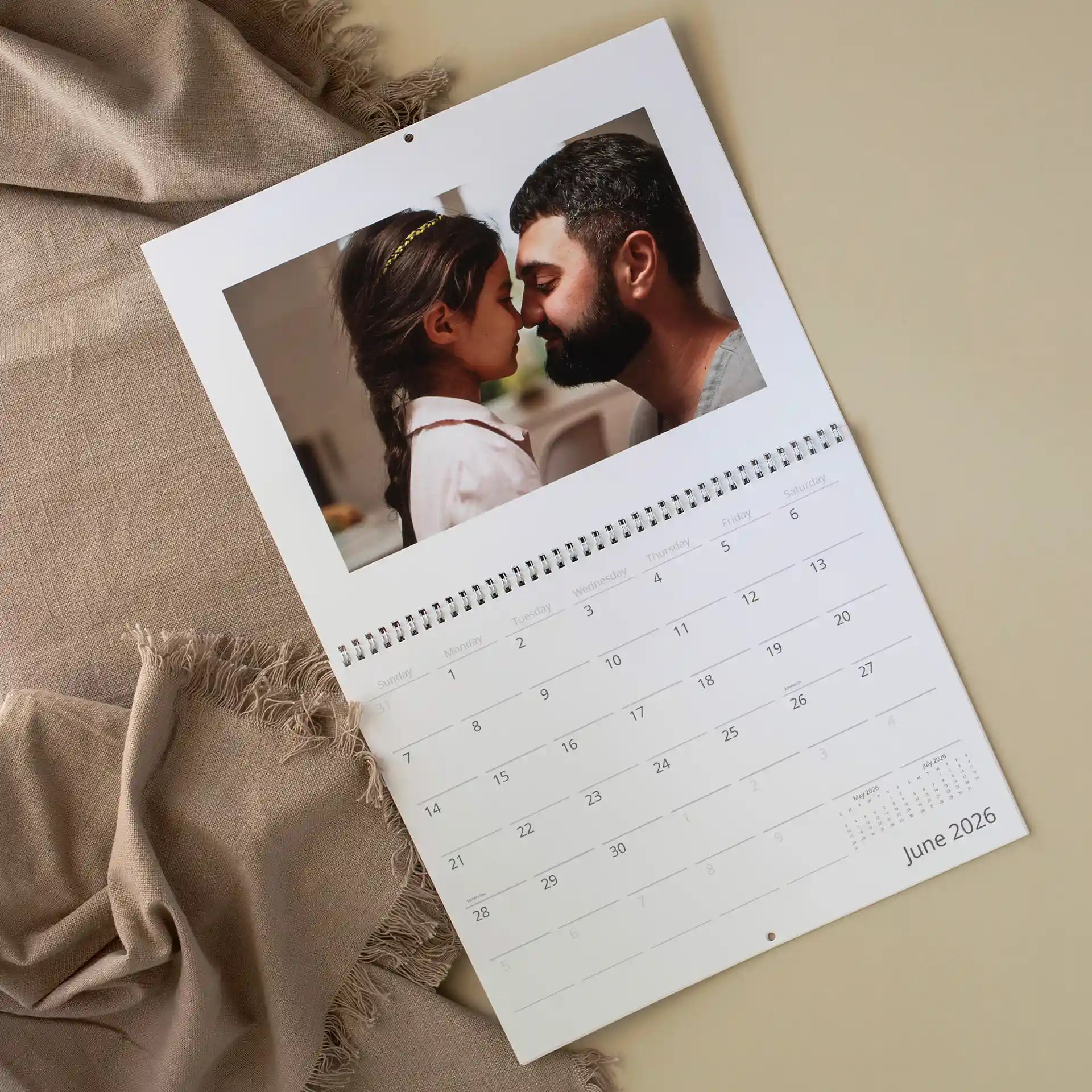 Custom wall calendar showing June 2026 resting on a linen surface, featuring a tender photo of a father and young daughter touching foreheads affectionately