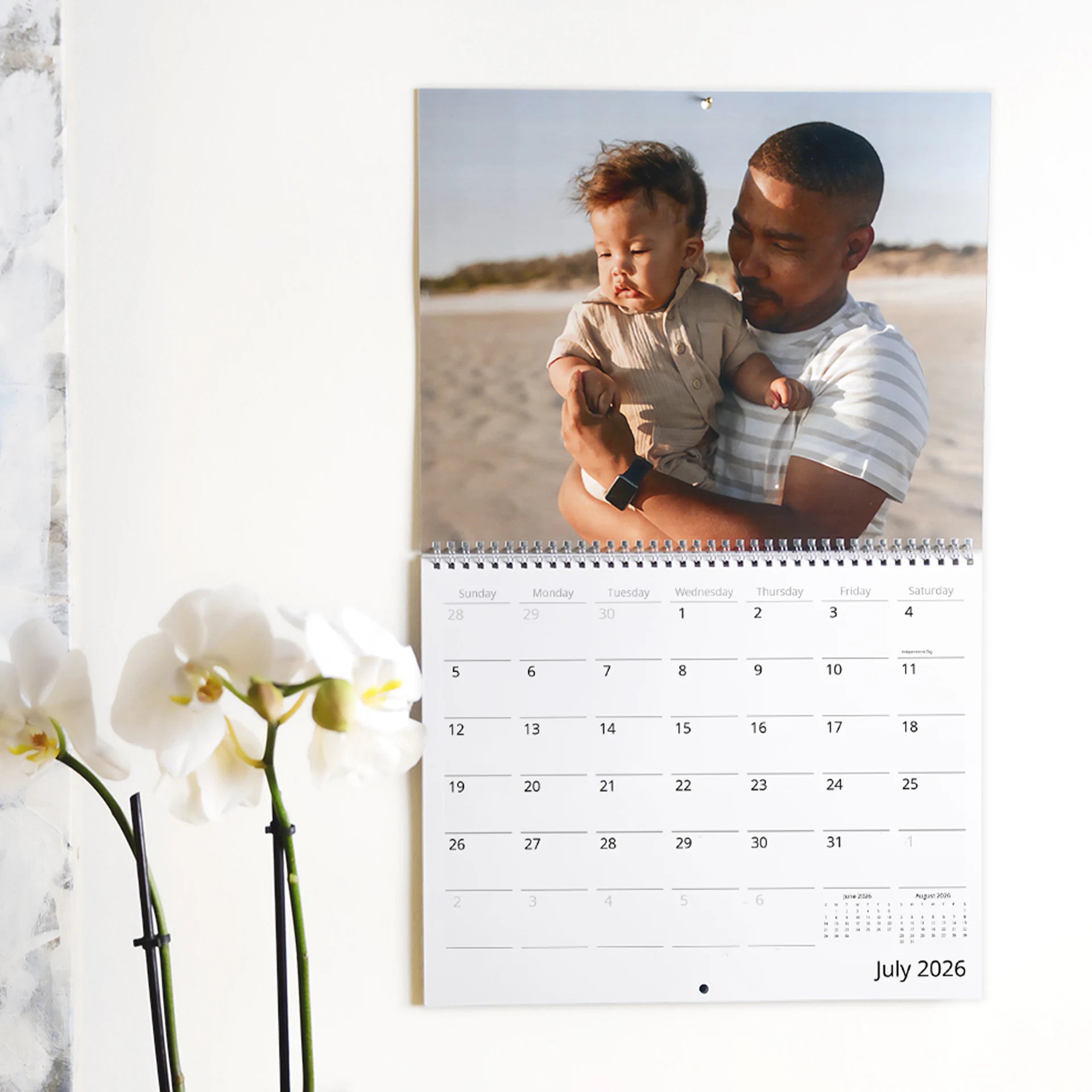 Wall calendar open to July 2026 with photo of adult holding baby on beach, displayed on wall next to white orchid.