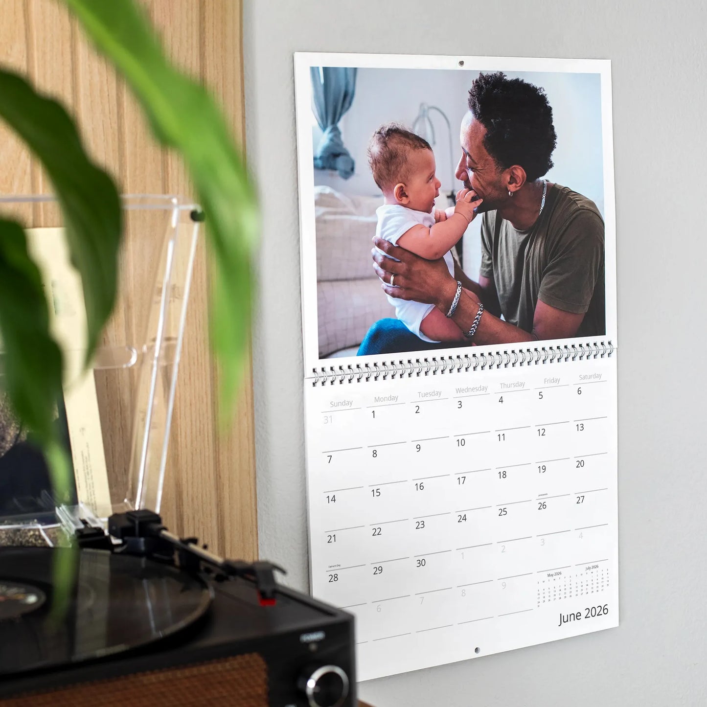 Custom wall calendar showing June 2026 hanging on a wall beside a record player and houseplant, featuring a photo of a father smiling as he holds his newborn baby indoors