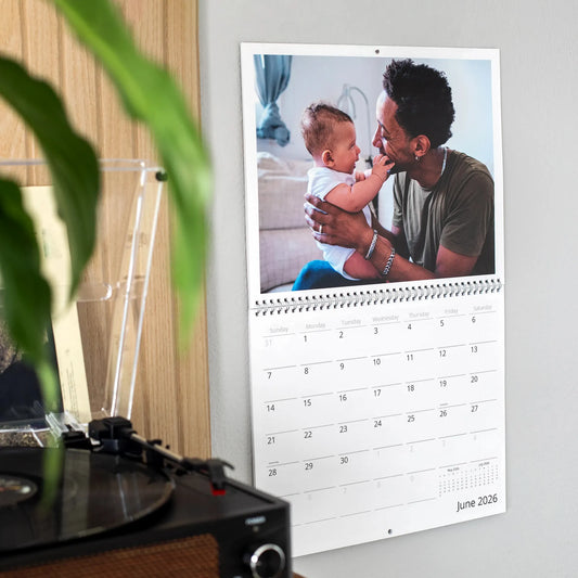 Custom wall calendar showing June 2026 hanging on a wall beside a record player and houseplant, featuring a photo of a father smiling as he holds his newborn baby indoors