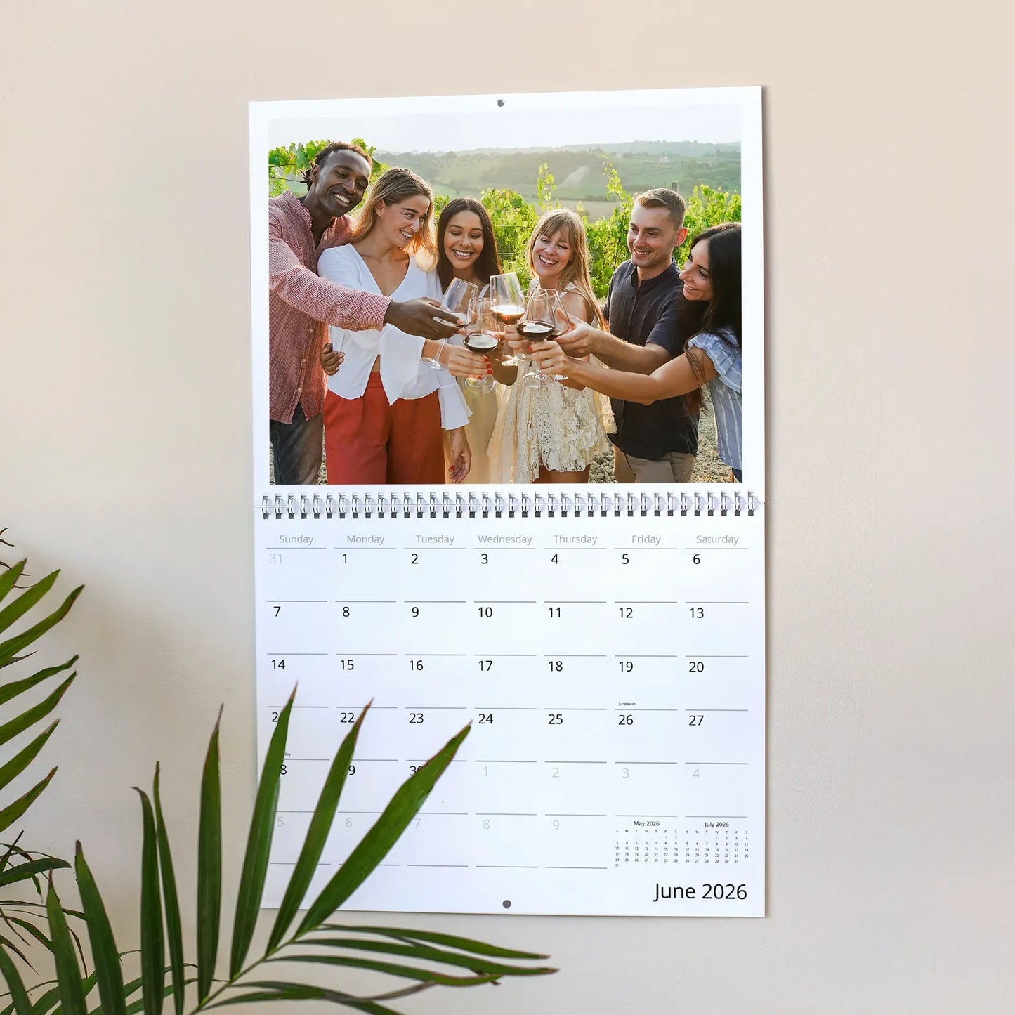 Wall calendar open to June 2026 showing group of adults toasting with wine glasses outdoors, hanging on wall with plant leaves visible.