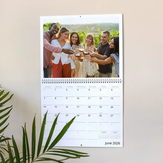 Wall calendar open to June 2026 showing group of adults toasting with wine glasses outdoors, hanging on wall with plant leaves visible.