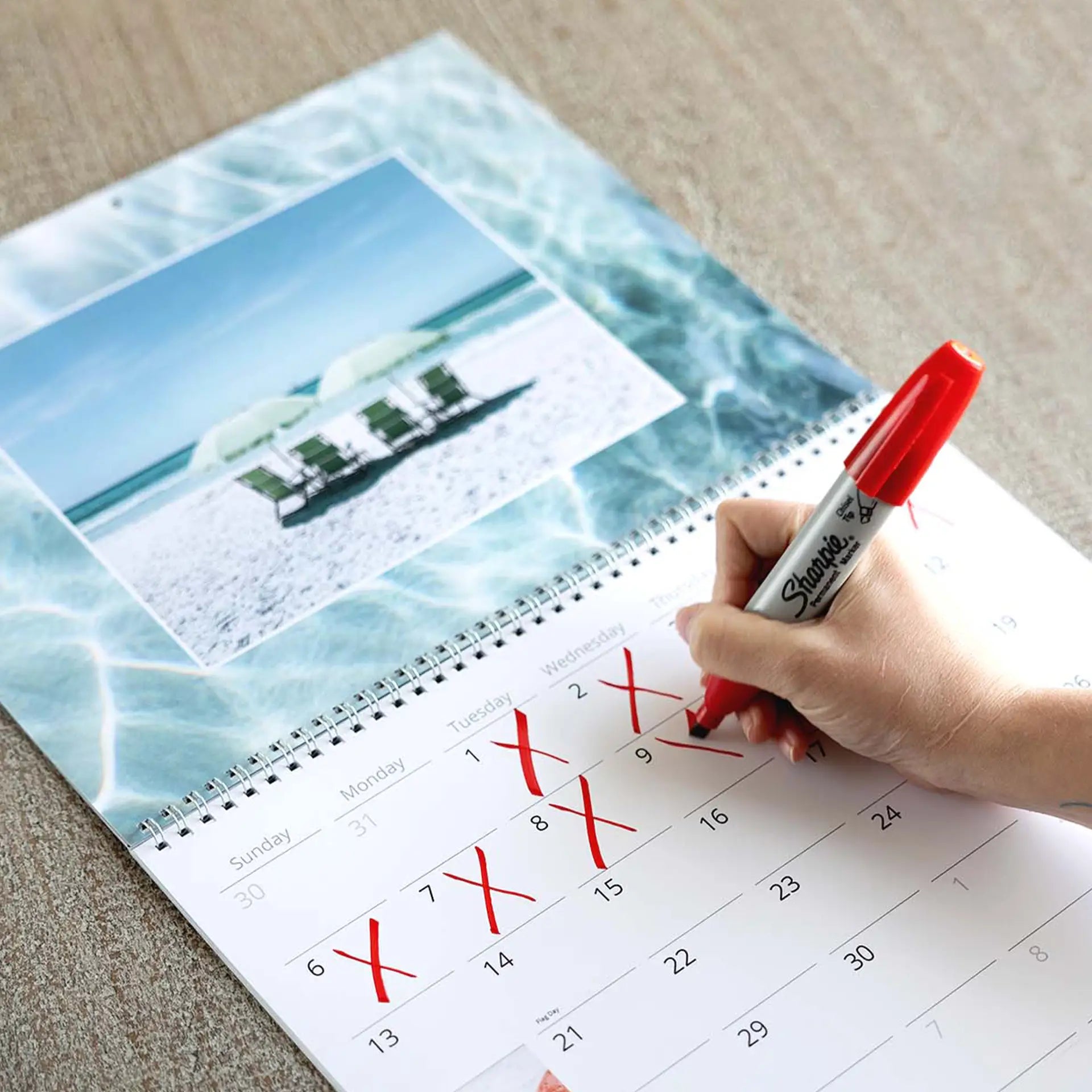 Wall calendar close-up with hand marking dates using red marker, beach photo at top and monthly grid visible.