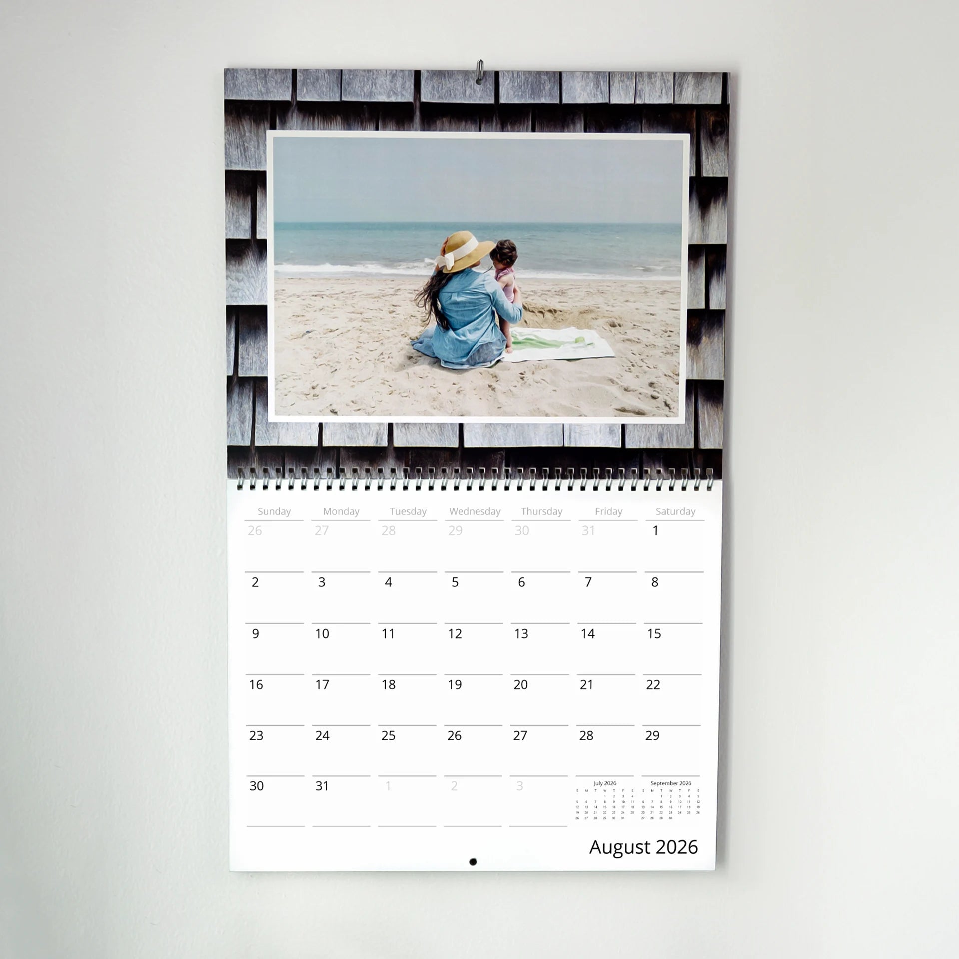 Wall calendar open to August 2026 with photo of adult and child sitting on beach, hanging against wood shingle background.
