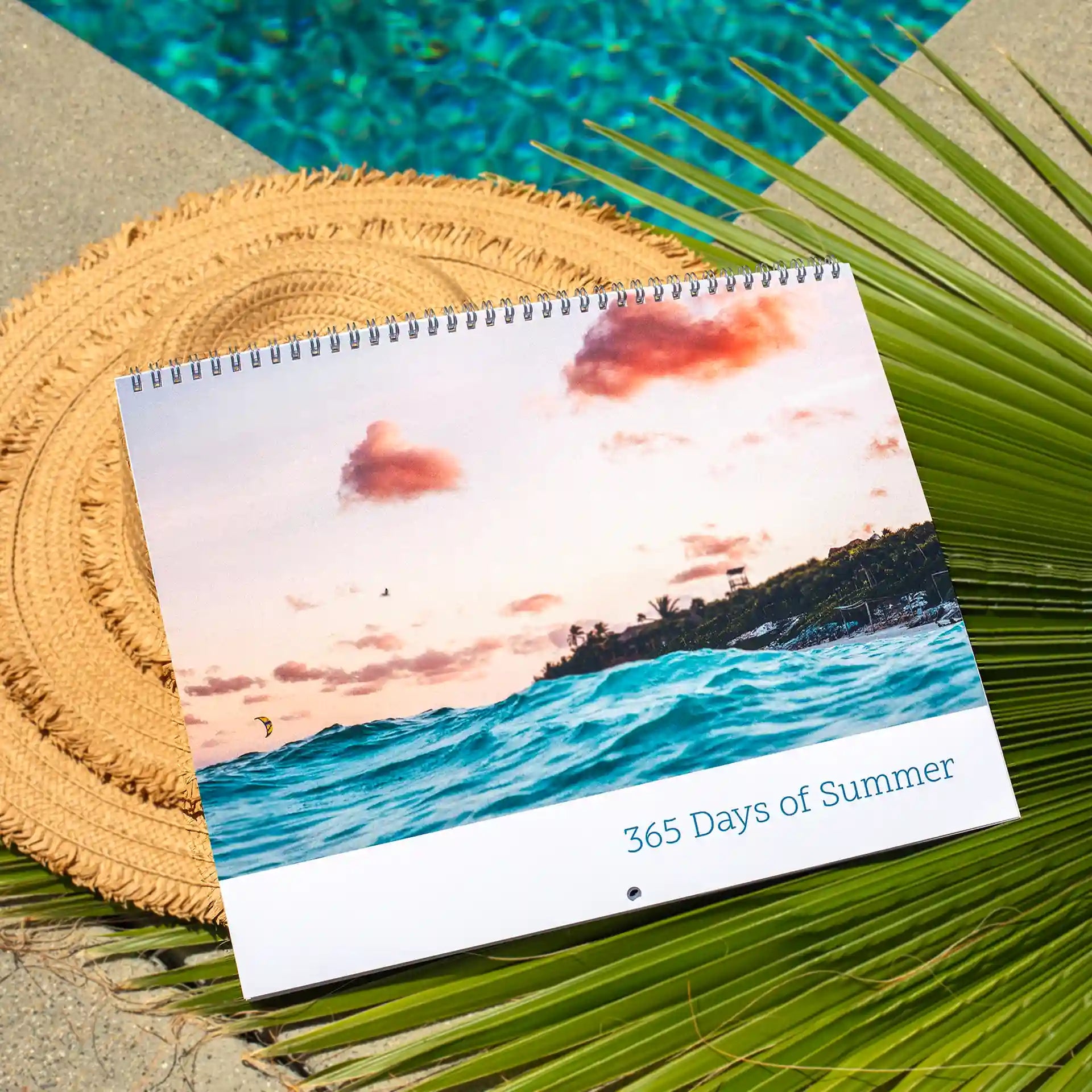 Wall calendar cover showing ocean waves at sunset with pink clouds, text reads “365 Days of Summer,” placed on woven mat with palm leaves.