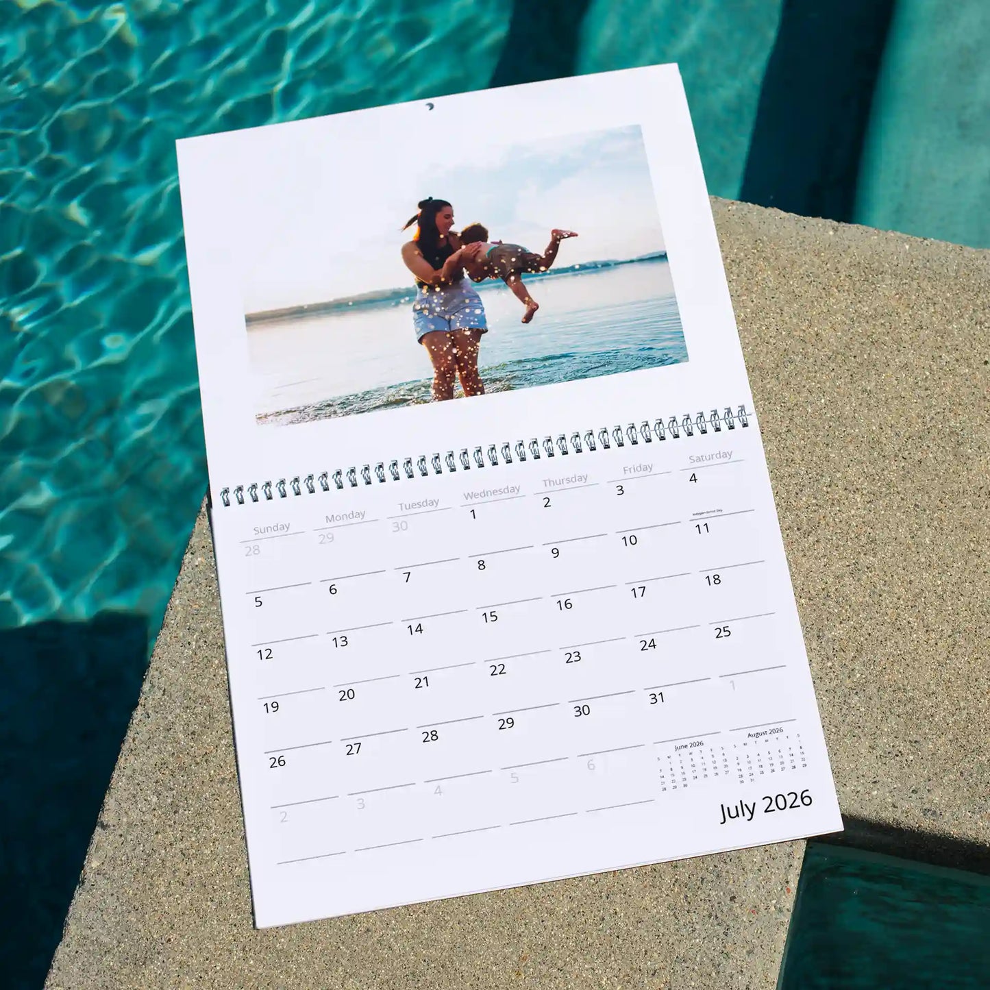 Wall calendar open to July 2026 with photo of adult lifting child in water, placed near pool edge with visible monthly grid.