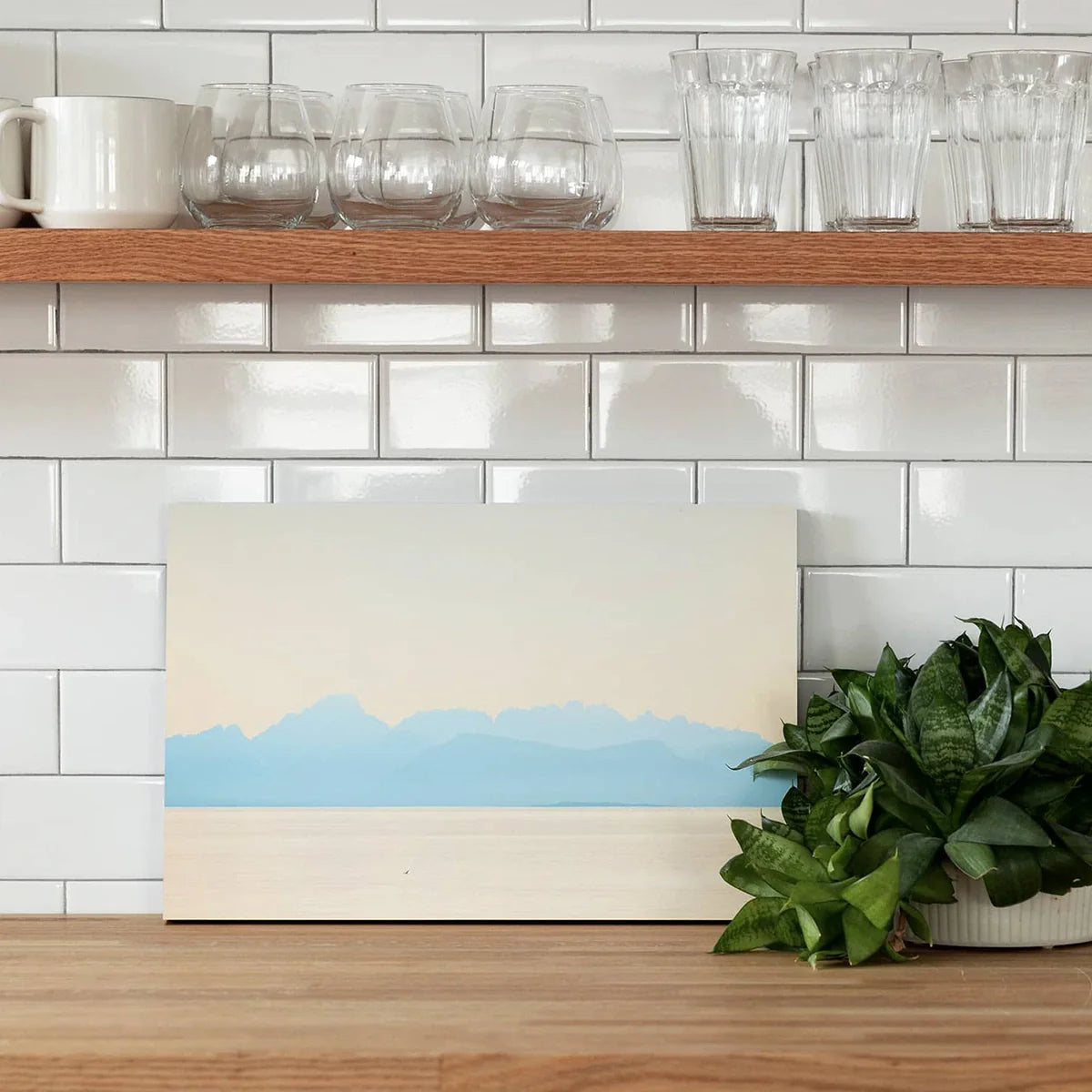 Mounted landscape print of soft blue mountains displayed on a kitchen counter with plants and glassware.
