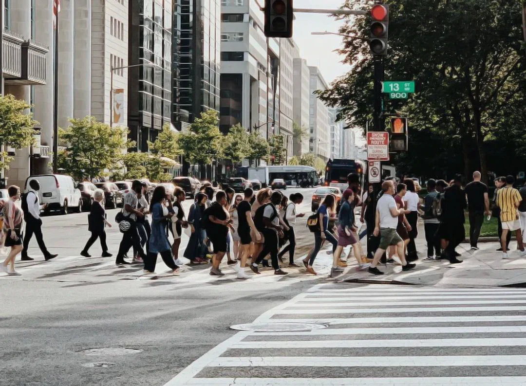 Crowd of pedestrians crossing a busy city intersection at a crosswalk between tall buildings during daytime with traffic lights and cars nearby.