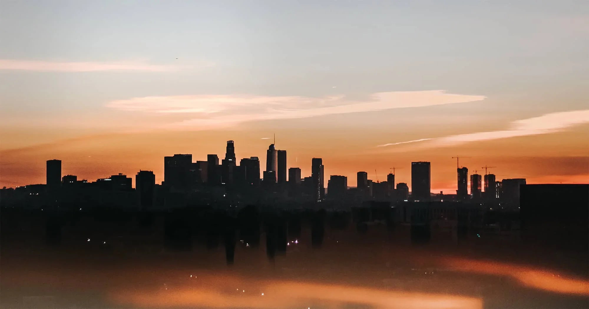 City skyline silhouette at sunset with tall buildings against an orange sky and low fog or haze covering the foreground.