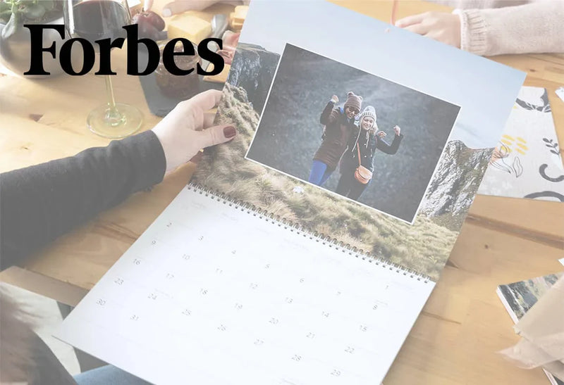 Person holding a custom photo wall calendar on a table with a printed image of two people hiking outdoors, with the Forbes logo.