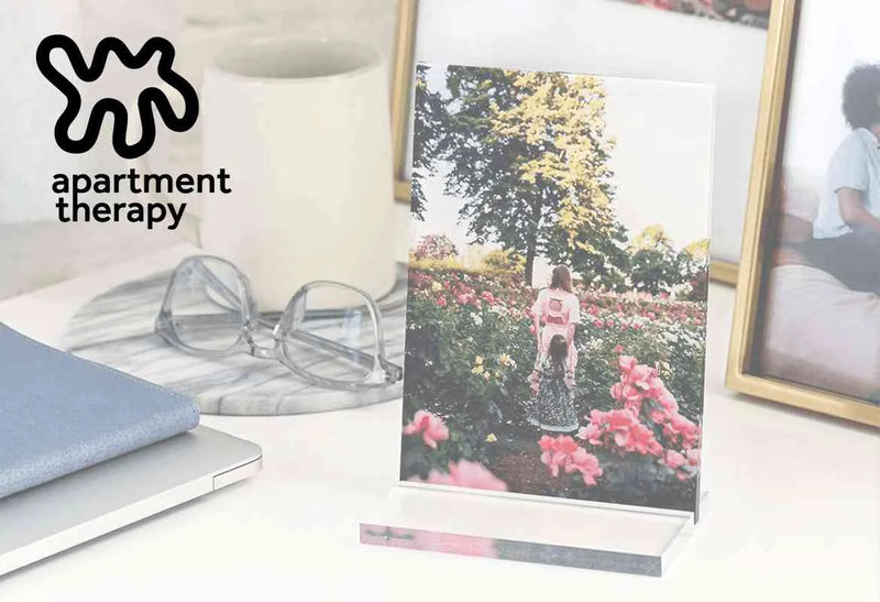 Desk setup with an acrylic desk print of a child walking through a flower garden next to glasses and a laptop, with the Apartment Therapy logo.