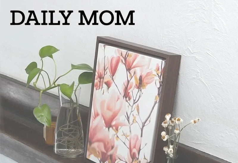 Framed canvas botanical art print with pink magnolia flowers displayed on a wooden shelf beside glass vases and a green plant, with “Daily Mom” text.