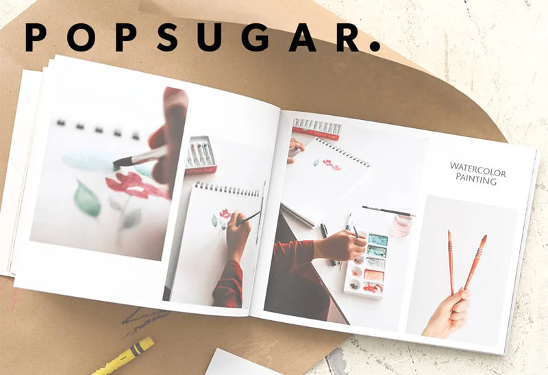 Open photo book displaying watercolor painting process images and art supplies on a craft table with paper and crayons, with “POPSUGAR” logo overlay.