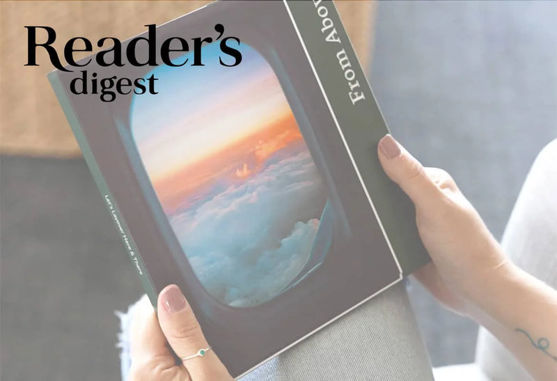 Hands holding a hardcover photo book with a window-view image of clouds and sunset on the cover, with “Reader’s Digest” text overlay.