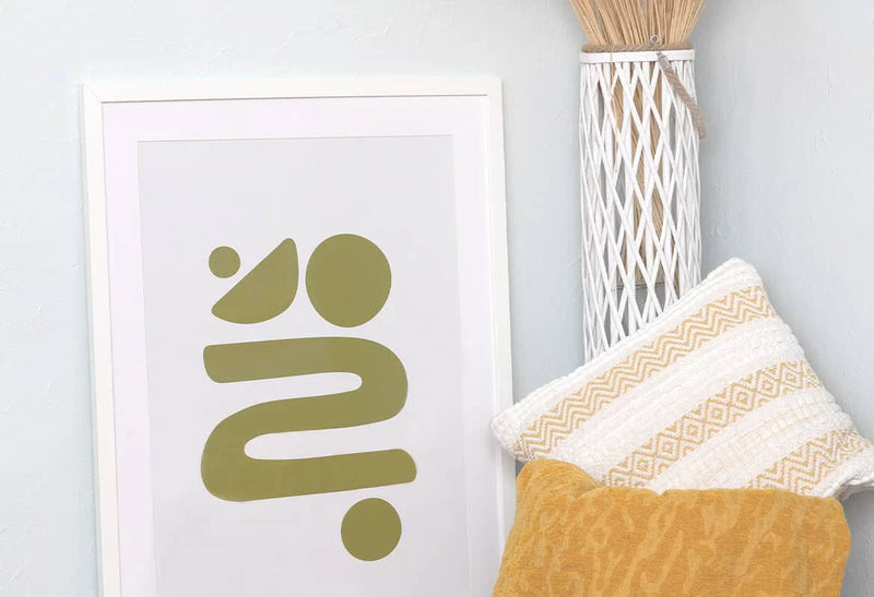 Framed minimalist abstract art print with olive green shapes leaning against a wall beside decorative pillows and a tall woven vase in a home interior.