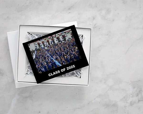 Hardcover photo book with black cover reading “Class of 2025,” displayed inside open gift box with tissue paper on marble surface.