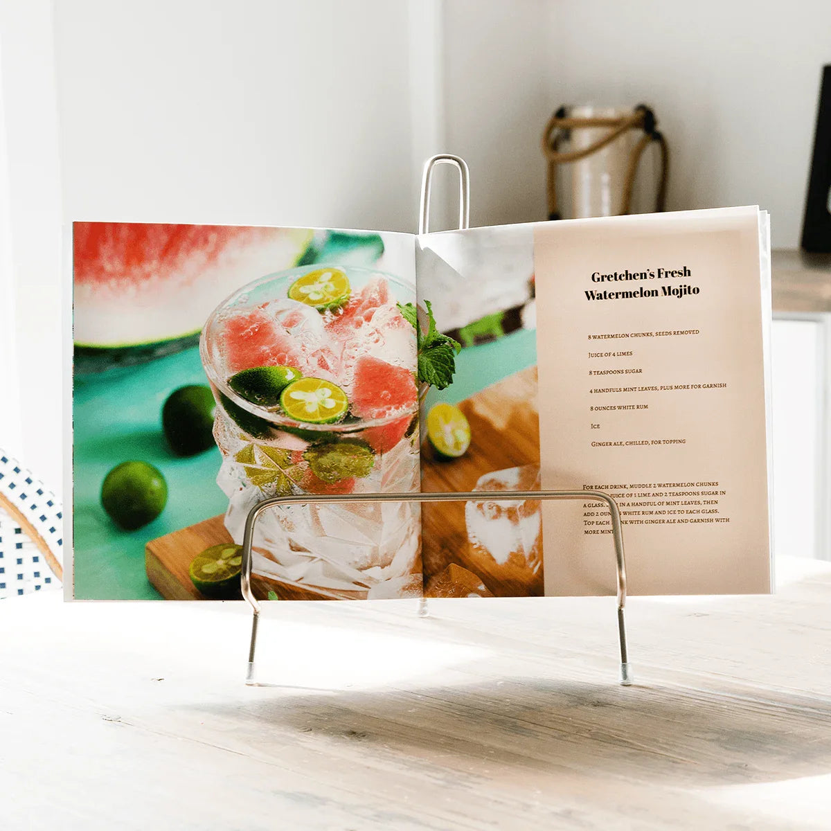 Open softcover photo book on a metal display stand showing a full-bleed cocktail image and recipe text on the opposite page.
