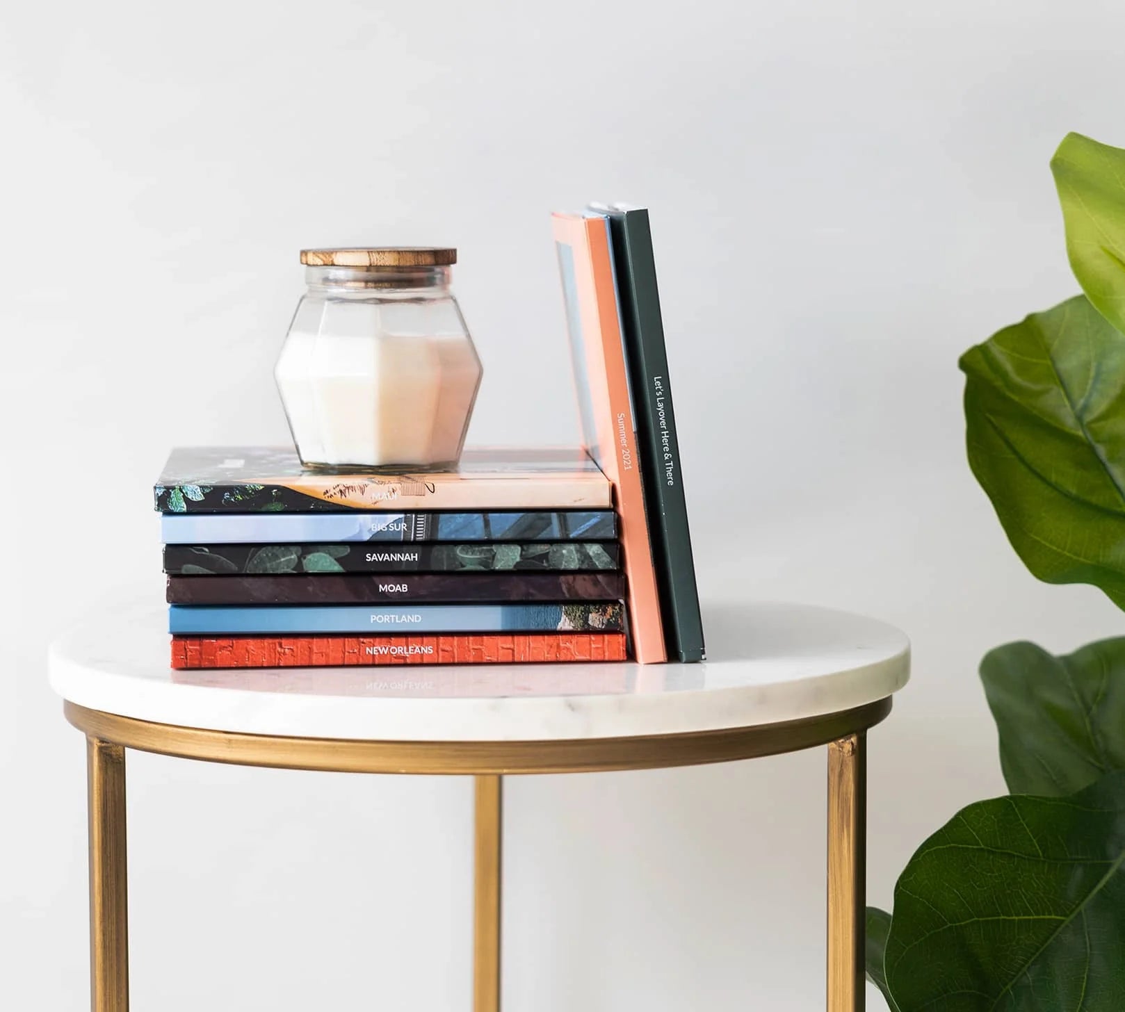 Stack of colorful hardcover photo books arranged on a round marble-top side table with gold legs, topped with a glass jar candle, and a leafy plant in the background.
