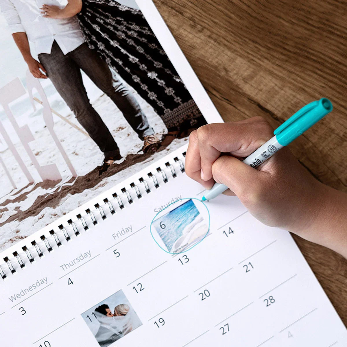 Close-up of a spiral-bound wall calendar with photo collage at top and monthly grid below, as a hand circles a beach photo date with a teal marker.
