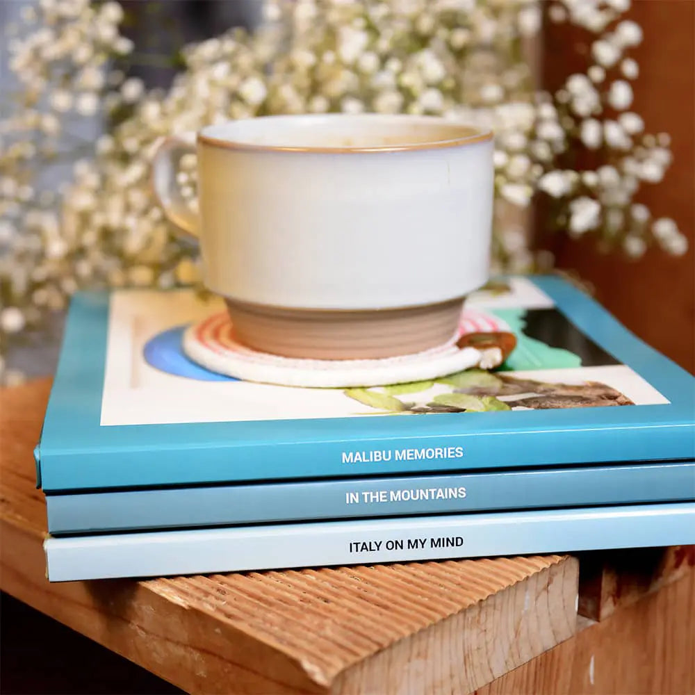 Stacked photo books with custom-printed spines on a wooden table, topped with a ceramic mug, highlighting personalized titles and clean design.