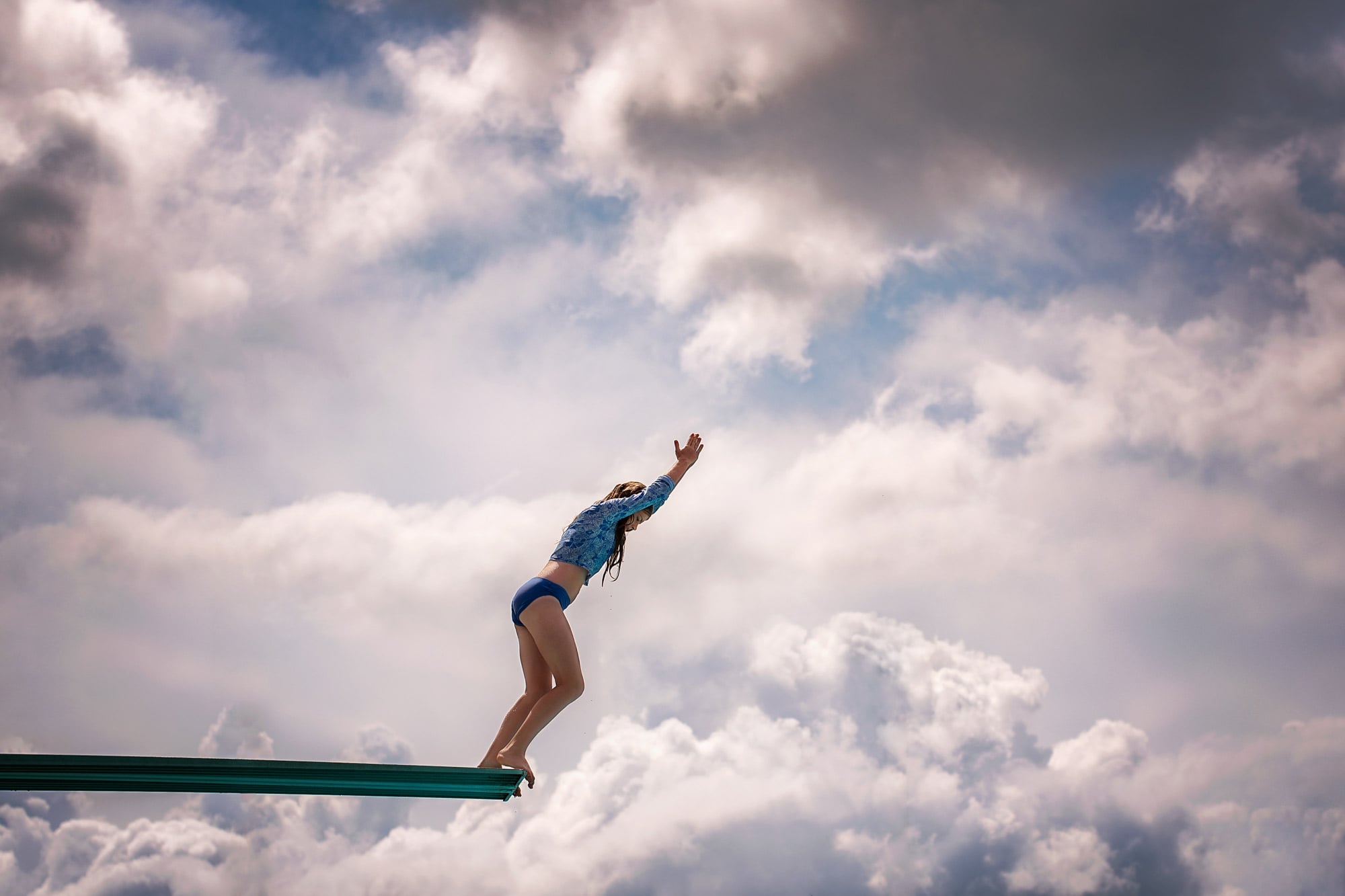 girl in diving position on diving board with blue sky and clouds in background 