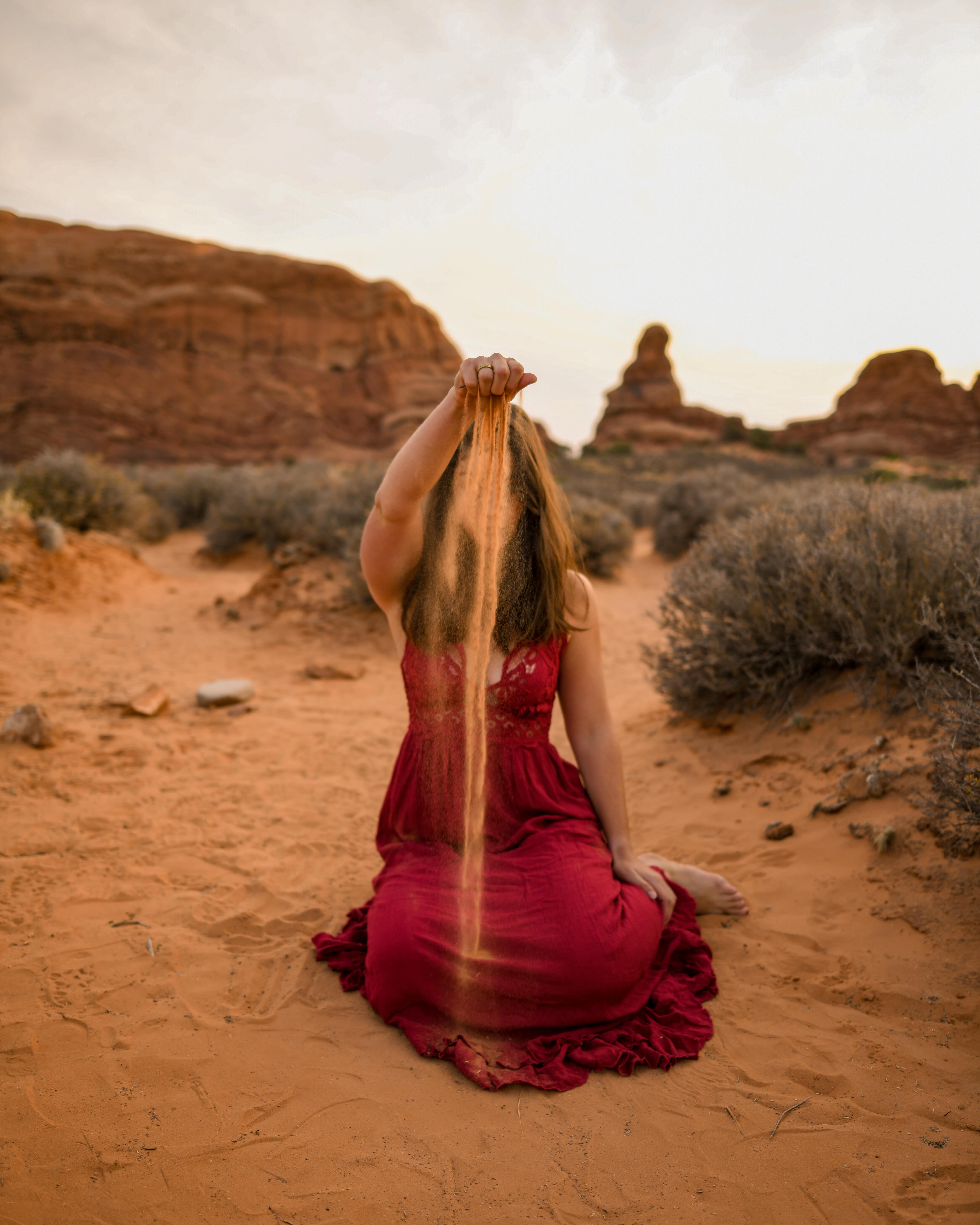 Girl in red dress sitting in sand and sprinkling sand infront of her face