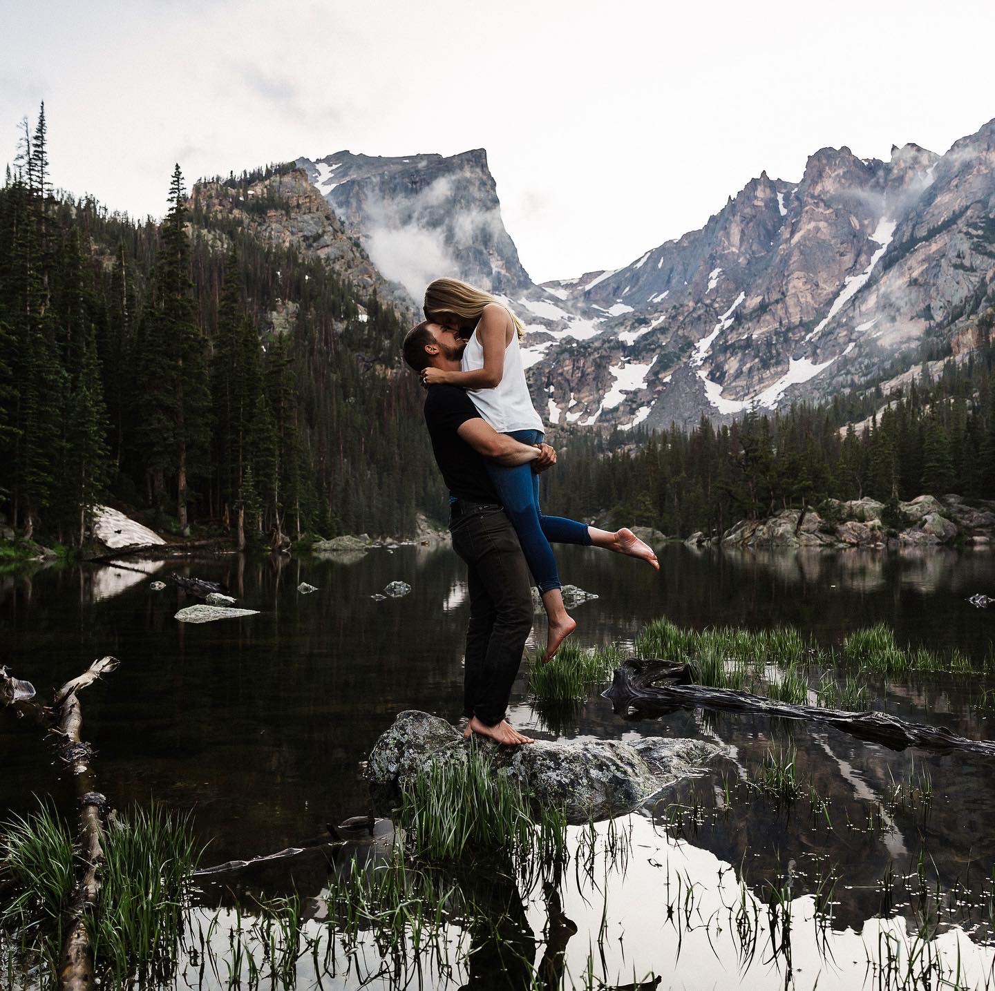 man standing on a rock in the water picking up woman with mountain landscape 